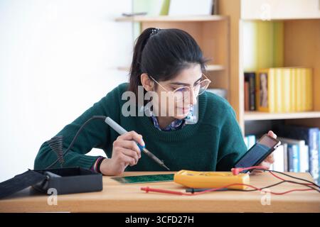 Young woman using a soldering iron and multimeter at a desk with bookshelves in the background. Germany Stock Photo