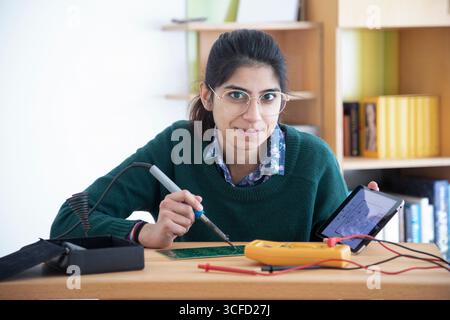 A woman in glasses soldering with a circuit board and holding a tablet at a desk. Germany Stock Photo