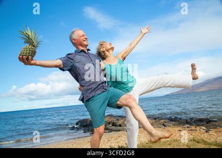 Senior couple balancing on one leg by the beach, sharing a playful moment with a pineapple. Kepaniwai Park, Wailuku, Hawaii Stock Photo