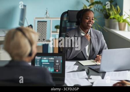 Young adult Black woman wearing headset smiling while participating in online meeting at office desk, sitting with notebook and laptop, Caucasian colleague working in foreground Stock Photo