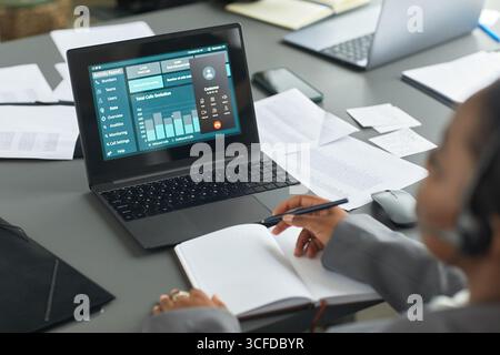 Black woman working at desk analyzing business data on laptop screen while taking notes in notebook, wearing headset, surrounded by documents, focusing on financial performance metrics Stock Photo