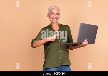 Smiling mature woman using a laptop while gesturing confidently against a beige backdrop, showcasing casual style and positive energy Stock Photo