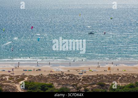 A vibrant telephoto view of a bustling sandy beach on a sunny day, with kite surfers on the deep blue ocean, framed by a shoreline Stock Photo