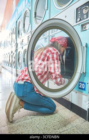Woman wearing bandana checking inside open tumble dryer at laundromat during laundry routine Stock Photo