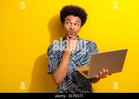 Young man with afro hairstyle holding a laptop thoughtfully in front of a yellow background, wearing a stylish patterned shirt Stock Photo