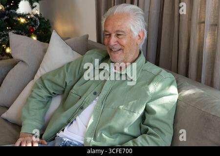 Senior man sitting on gray sofa in living room with Christmas tree, tinsel and cushions Stock Photo