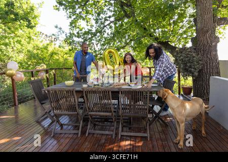 Diverse friends arranging party setup on wooden deck with dining table gold '0' balloon, dog Stock Photo