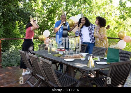 Diverse friends wearing party hats blowing noisemakers around table on deck with cake, balloons Stock Photo