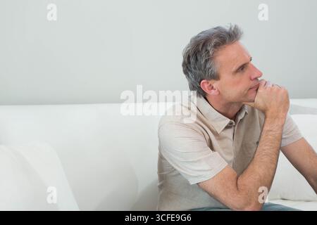 Middle-aged man sitting on white sofa leaning elbow on knee in living room, copy space Stock Photo