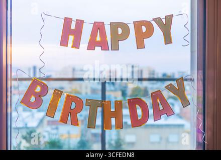 Close up of Happy Birthday banner hanging on window glass with blurred cityscape in the background Stock Photo