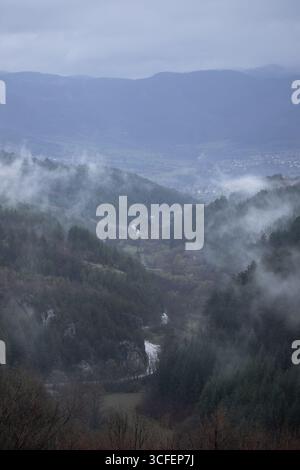 A foggy mountain valley stretches into the distance, where a winding river and narrow road snake through the dense pine forest. Stock Photo
