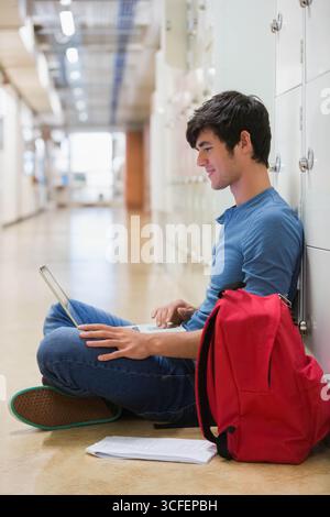 Male student sitting in hallway studying on laptop next to red backpack and papers, copy space Stock Photo