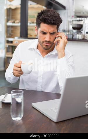 Businessman using smartphone for phone call while sipping coffee at coffee shop table with laptop Stock Photo