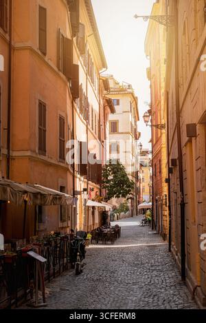 Narrow cobblestone street in Trastevere, Rome, Italy, with cafes, tables, warm sunset light and colorful buildings Stock Photo