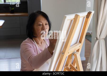 Senior asian woman in white apron using paintbrush painting canvas at easel in home art space Stock Photo
