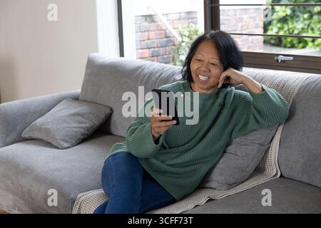 Senior asian woman using smartphone while reclining on couch pillows in living room near window Stock Photo