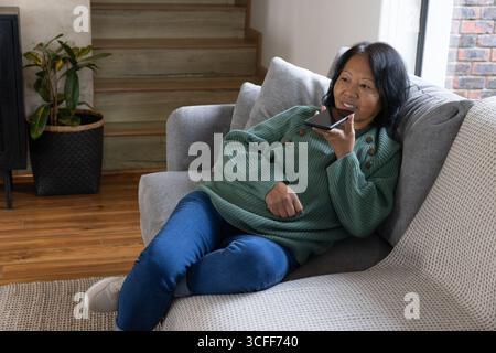 Senior asian woman relaxing on sofa at home talking into smartphone by staircase and plant Stock Photo