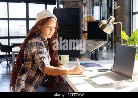 African American woman writing code notes while glancing at monitor and laptop at office desk Stock Photo