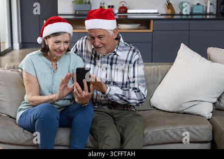 Senior couple wearing Santa hats using smartphone on grey leather couch in open kitchen at home Stock Photo