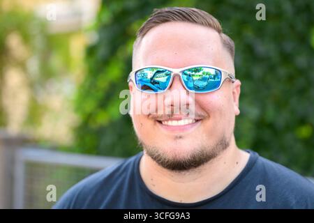 A young man wearing white-framed mirrored sunglasses and a black T-shirt smiles broadly in a close-up outdoor portrait. Stock Photo