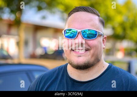 A young man wearing white-framed mirrored sunglasses and a black T-shirt smiles warmly while standing outdoors. Stock Photo