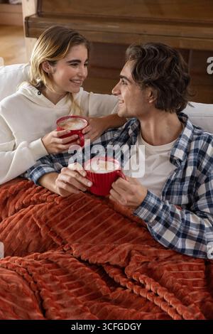 Couple sitting on sofa under red blanket holding coffee mugs in living room by wooden piano Stock Photo