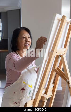 Senior asian woman in paint-stained apron painting canvas on wooden easel in home living area Stock Photo