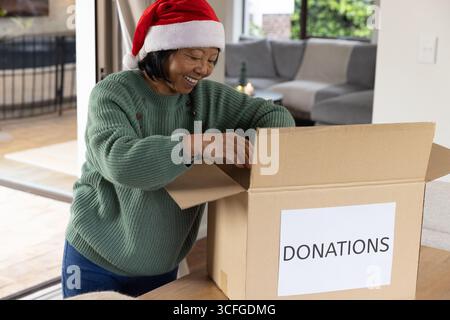 Senior Asian woman wearing Santa hat opening donations box in living room at table near sofa Stock Photo