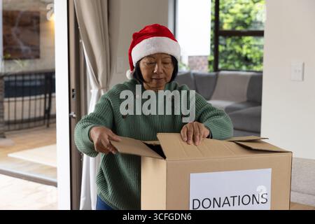 Asian woman opening cardboard donation box in living room wearing Santa hat and green sweater Stock Photo