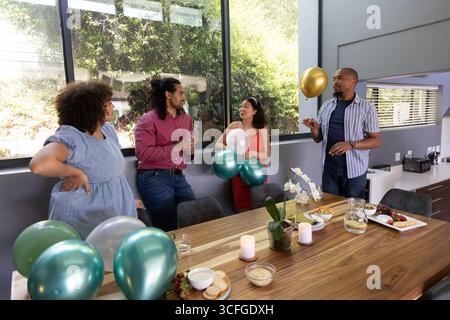 Diverse friends gathering around long wooden table in modern dining room with balloons Stock Photo
