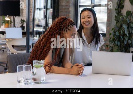 Diverse female coworkers collaborating over laptop at table in office with pitcher of lime water Stock Photo