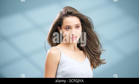 Young woman with radiant skin and long, flowing hair poses elegantly against a soft blue backdrop, showcasing her natural beauty and confidence in a s Stock Photo