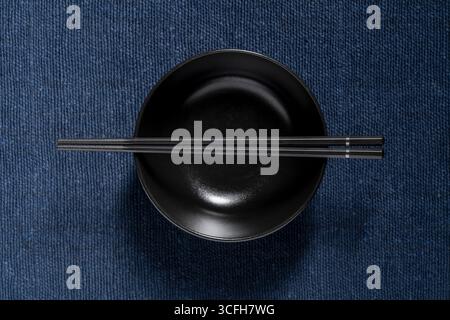 A pair of chopsticks lying across an empty black soup bowl on blue fabric background, close up, top view Stock Photo