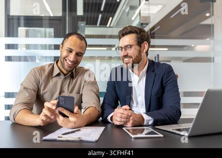 Two business professionals smiling while discussing work using a smartphone and tablet at their workplace. The modern office environment emphasizes teamwork, collaboration, and technology. Stock Photo