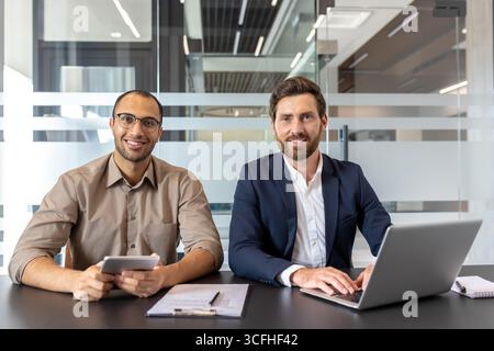 Two business professionals are sitting together, discussing plans and sharing ideas. They convey collaboration, teamwork, and focus while using modern technology in a contemporary office setting. Stock Photo
