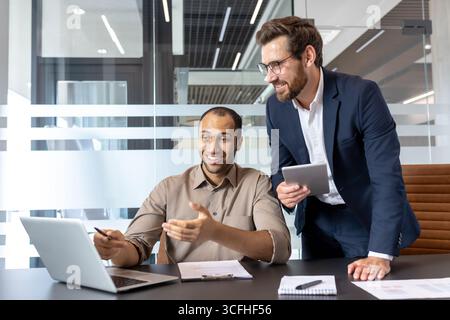 Two professionals engaged in a collaborative discussion with a laptop, highlighting teamwork and productivity in a modern office setting. Stock Photo