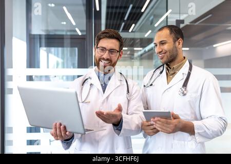 Two professional doctors in white coats discussing medical information on a laptop and tablet. Collaboration in a modern hospital environment showcasing teamwork and technology integration. Stock Photo