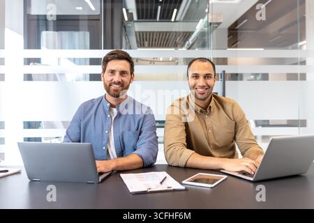 Two colleagues sitting side by side with laptops in a bright and contemporary workspace, smiling and engaging, showcasing collaboration and teamwork in a professional environment. Stock Photo