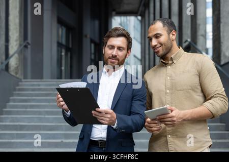 Two professional men discuss work outside an office building, holding documents and tablet, showcasing collaboration. Stock Photo
