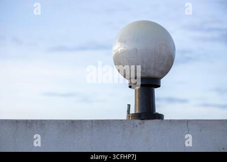 A round white glass lamp, mounted on a black steel pole attached to a wall on a house rooftop, with a blurred sky in the background. Stock Photo