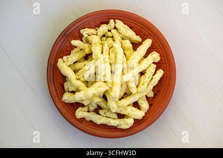 Tasty corn stick chips in a clay plate on a light wooden surface. Kurkure American-style cream and onion flavor chips, a delicious savory snack food. Stock Photo