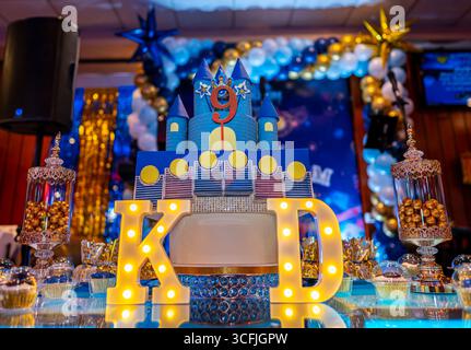 Blue castle cake with number 9 topper, illuminated letters K and D, golden candies, and festive decoration at a celebration event. Stock Photo