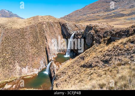 Waterfall plunging from rocky cliffs into emerald pools in the Peruvian Andes, surrounded by dry grasslands and rugged highland terrain. Stock Photo