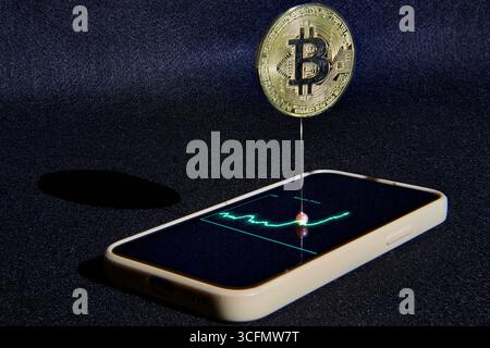 A physical Bitcoin emerges from a phone's stock chart, balanced on a pin. A powerful concept for cryptocurrency bubbles, market volatility, fintech in Stock Photo