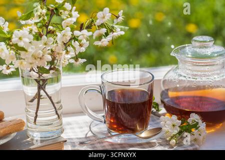 Delicious hot tea on the windowsill at home at summer day near garden, close up, copy space. Hot black tea in a glass teapot and cup Stock Photo