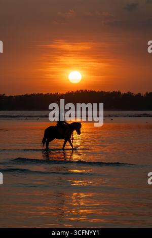 Dramatic silhouette of a rider on horseback wading through shallow waters with a vivid orange sunset and cloudy sky over Gili Air, Indonesia. Stock Photo