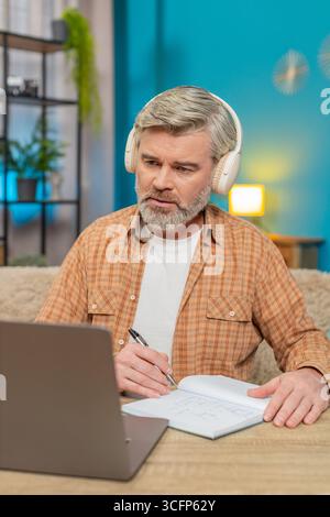 Caucasian mature man sitting at home table listening to podcast or online stream on laptop with headphones. Guy takes making notes into notebook, interested engaged in content, learning education Stock Photo