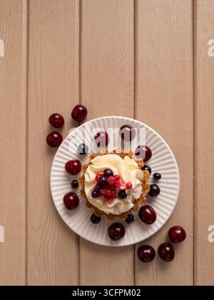 Close-up of a berry tart with whipped cream, topped with cherries, blueberries, and raspberries. Served on a white plate with extra cherries, all plac Stock Photo