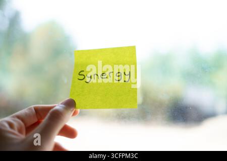 Hand holding a yellow sticky note with the word synergy written on it in front of a window Stock Photo