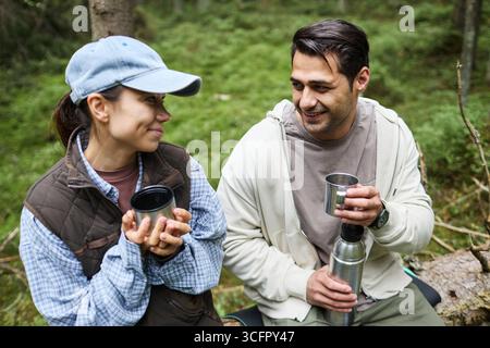 Young adult Caucasian man and young adult Caucasian woman sitting outdoors in forest, holding metal cups and thermos smiling at each other enjoying break during hike Stock Photo